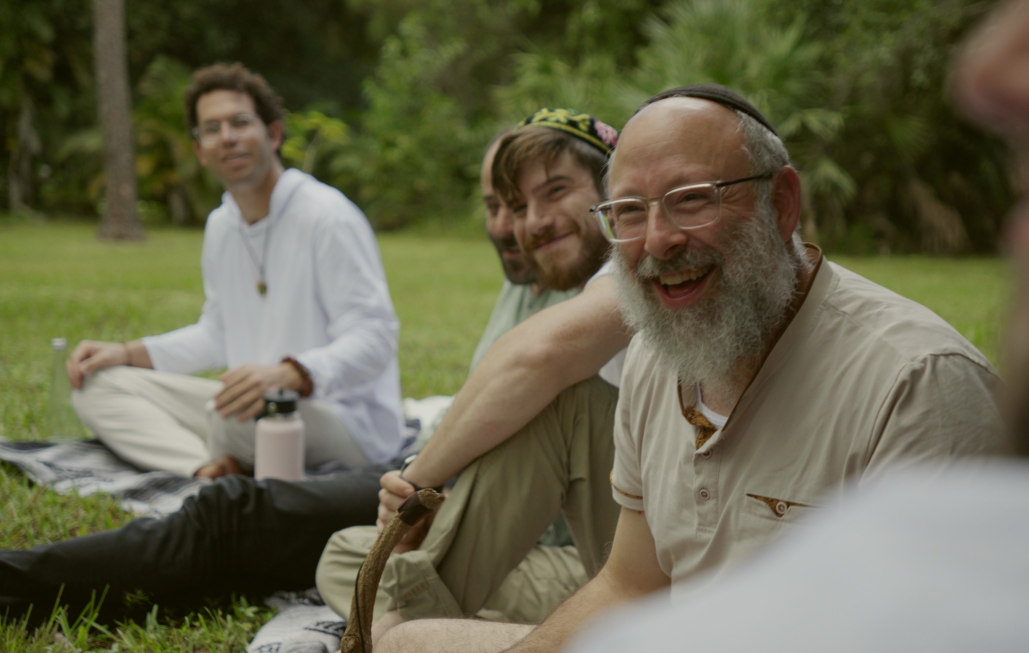 A group sitting on blankets on the grass in daylight, an older man laughing openly and the others smiling with him.