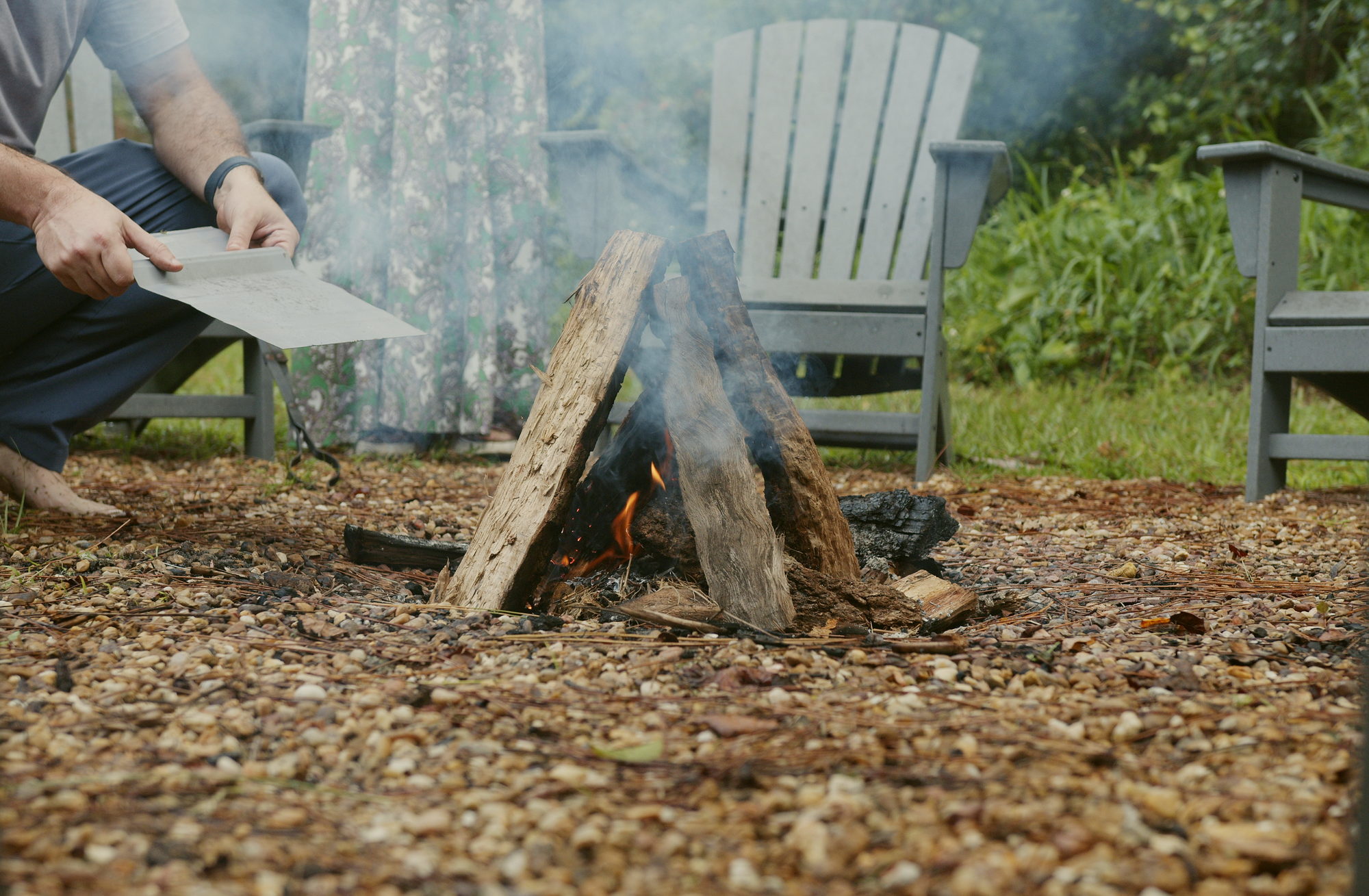 Hands fanning a small fire with a piece of card, smoke lifting across the gravel.