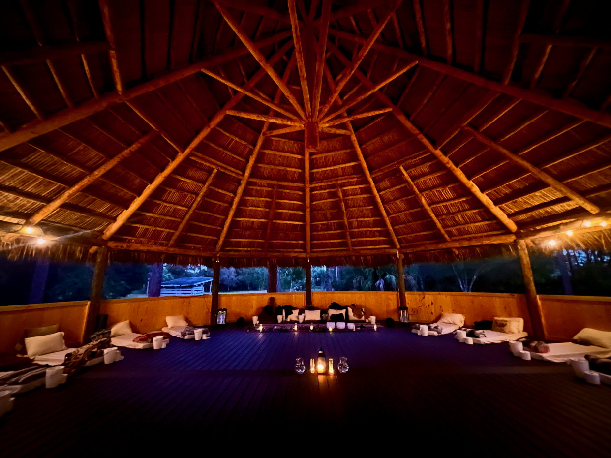 The maloka at dusk from inside: a high thatched roof on timber posts, open on every side to the trees, with mats and cushions laid in a circle and candles lit around them.