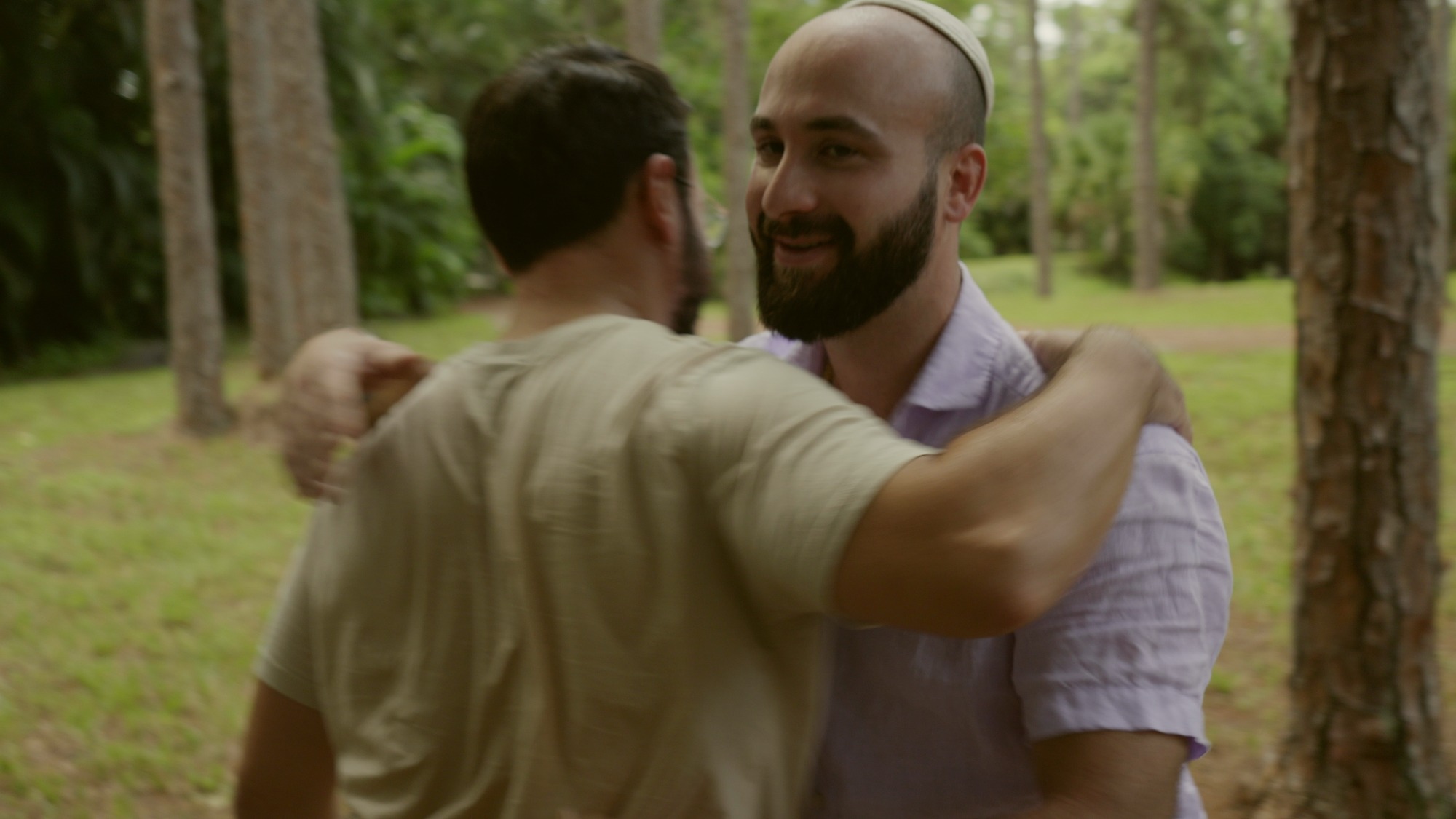 Two men meeting on the lawn among pine trunks, arms round each other's shoulders, one smiling at the other.