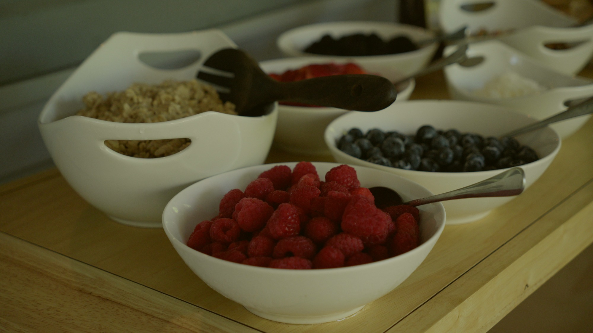 White bowls of raspberries, blueberries and oats set out on a wooden counter.