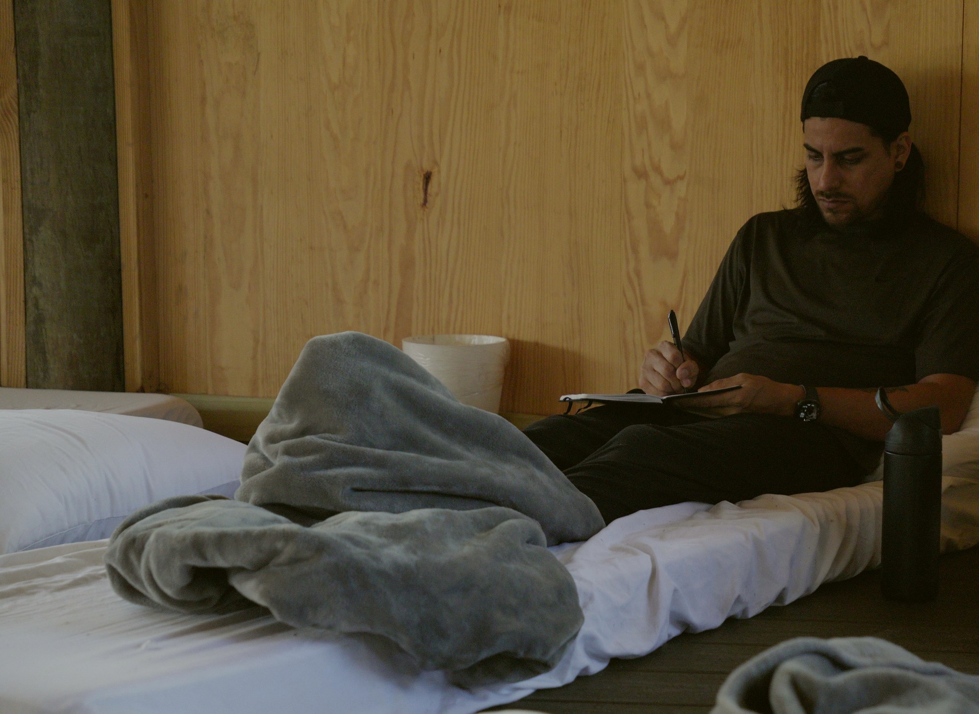 The shared sleeping space: bedding laid out on the floor against a timber wall, someone sitting up writing in a notebook.