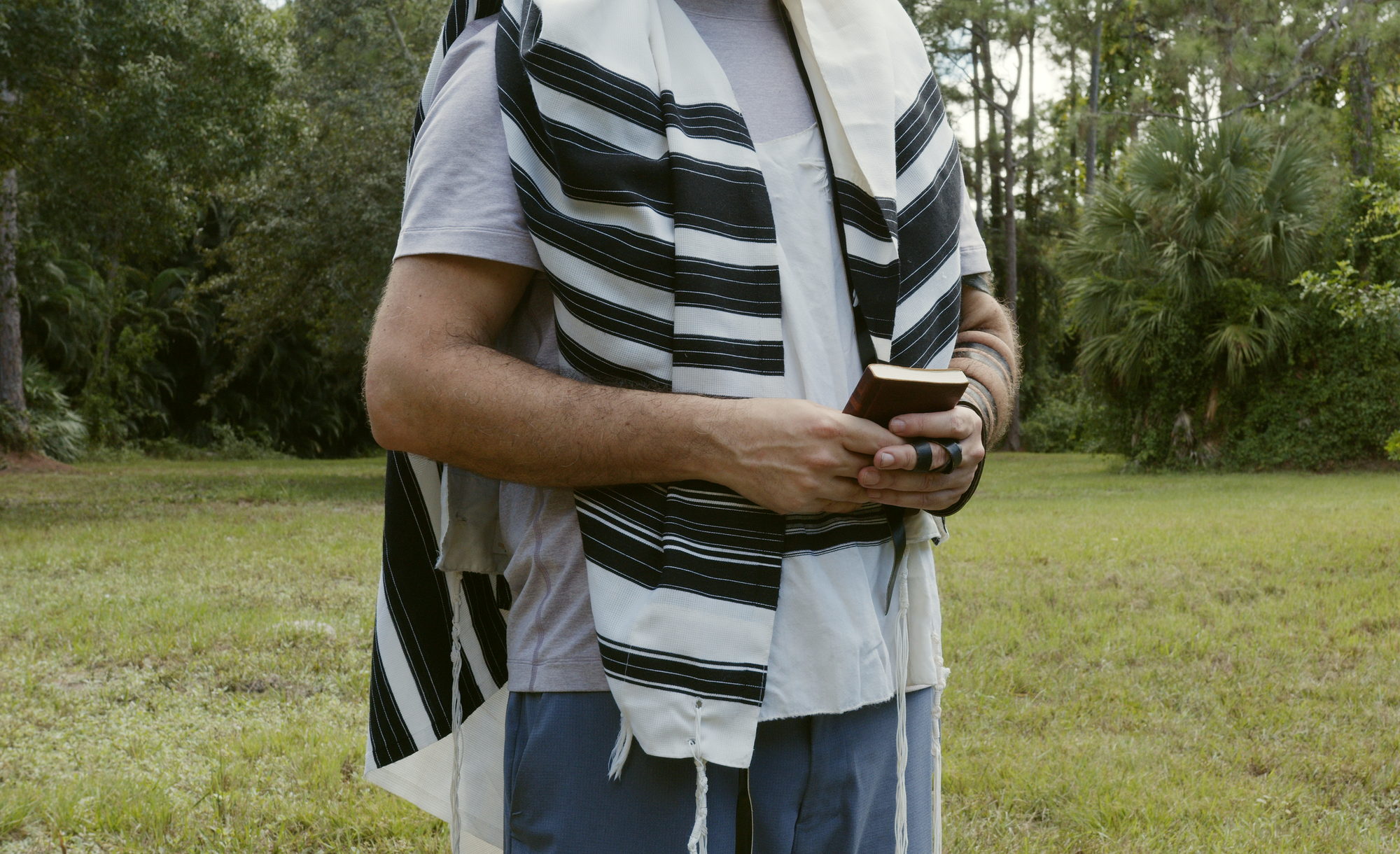 A man standing on the grass wrapped in a prayer shawl, holding a prayer book.