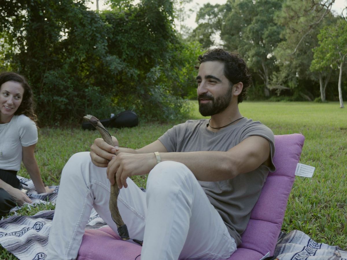 Two people sitting and talking on a blanket on the grass, trees behind them.